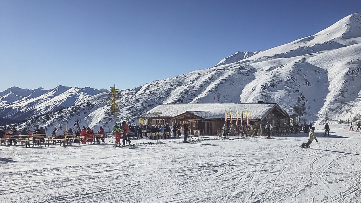 Blick aus der Ferne zur Skihütte Gruobenalp bei welcher Skifahrer und Snowboarder auf der Piste vorbei kurven. | © Davos Klosters Mountains