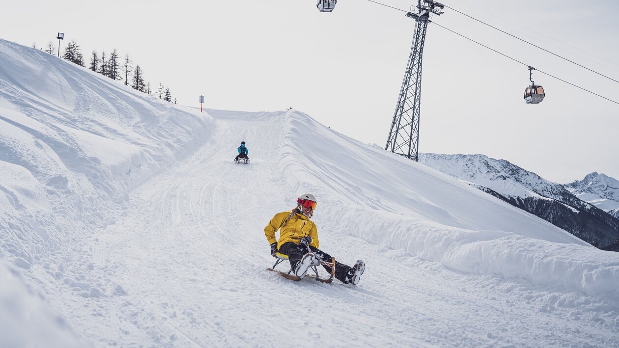 Zwei Freundinnen schlitteln auf der Schlittelstrecke Rinerhorn um eine der 24 Kurve mit der Rinerhornbahn im Hintergund. | © Davos Klosters Mountains